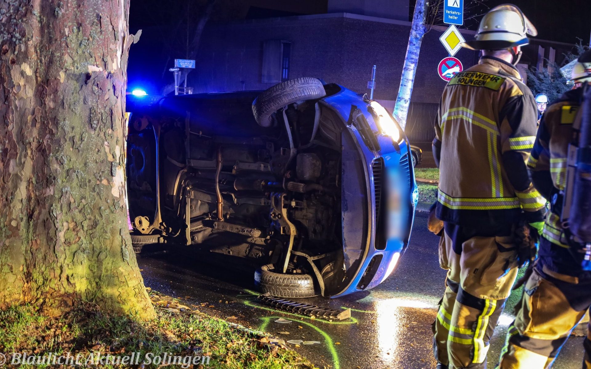 Alt Text: Einsatzkräfte der Feuerwehr bei einem Autounfall in Solingen, Fahrzeug auf der Seite neben einem Baum.